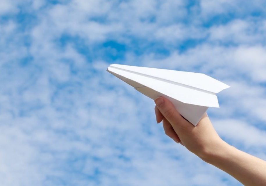 A hand holding a paper airplane aloft against a blue sky with clouds